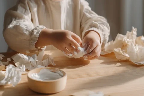 Kleine kinderhanden met een beetje witte verf erop bezig met knutselwerk aan een lichte houten tafel in ochtendlicht